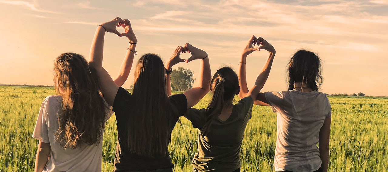 Groupe de jeunes femmes vues de dos dans un champ, elles font le signe du cœur avec leurs mains