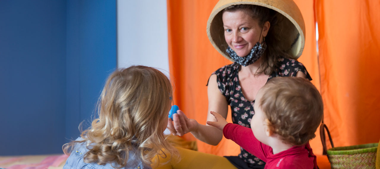 Une animatrice coiffée d’un chapeau improvisé (un grand saladier en bois) s’amuse avec deux enfants. Elle leur montre un objet bleu qui ressemble à un œuf.