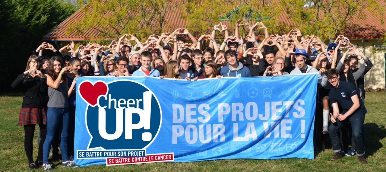 Portrait de groupe des équipes de l’association CHEER UP et des bénéficiaires. Ils soutiennent une banderole indiquant “Des projets pour la vie” devant eux. Ceux qui sont en arrière-plan font le signe du cœur. 