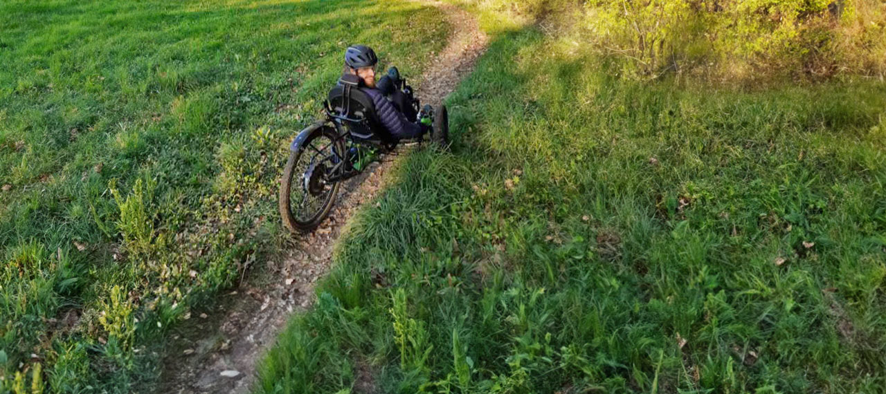 Un homme pratique le handbike (vélo modulable adapté aux handicaps de motricité) sur un chemin de terre dans un paysage verdoyant.