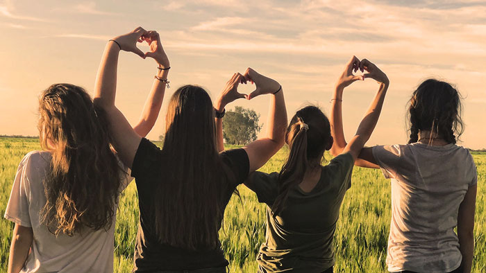 Groupe de jeunes femmes vues de dos dans un champ, elles font le signe du cœur avec leurs mains.