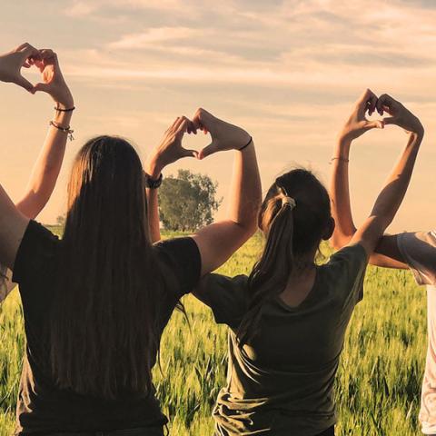 Groupe de jeunes femmes vues de dos dans un champ, elles font le signe du cœur avec leurs mains