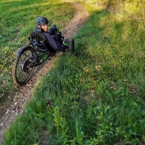 Un homme pratique le handbike (vélo modulable adapté aux handicaps de motricité) sur un chemin de terre dans un paysage verdoyant.