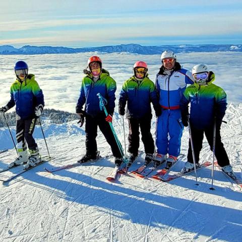Six skieurs, quatre adolescents et un moniteur, posent face caméra en haut d’une piste de ski ensoleillée. En arrière-plan la vallée est couverte d’une “mer de nuages”.